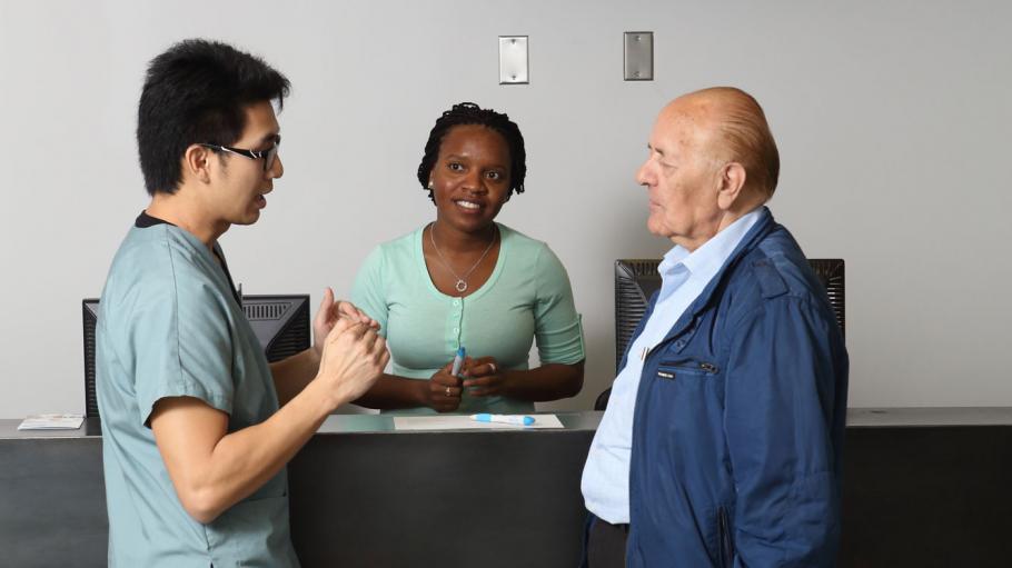 A female receptionist and a male Health Sciences student are helping an elderly visitor into the office at the receptionist desk. The elderly visitor is facing the student and having a conversation with him while the receptionist listens along.