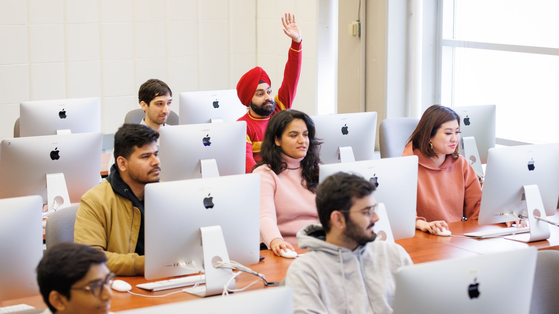 Students sit behind computers in rows in a classroom. One student has their hand raised.