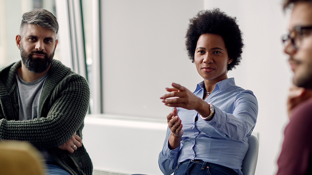 iStock Image — Small group discussion in a meeting room, with one person gesturing while speaking and others seated nearby.
