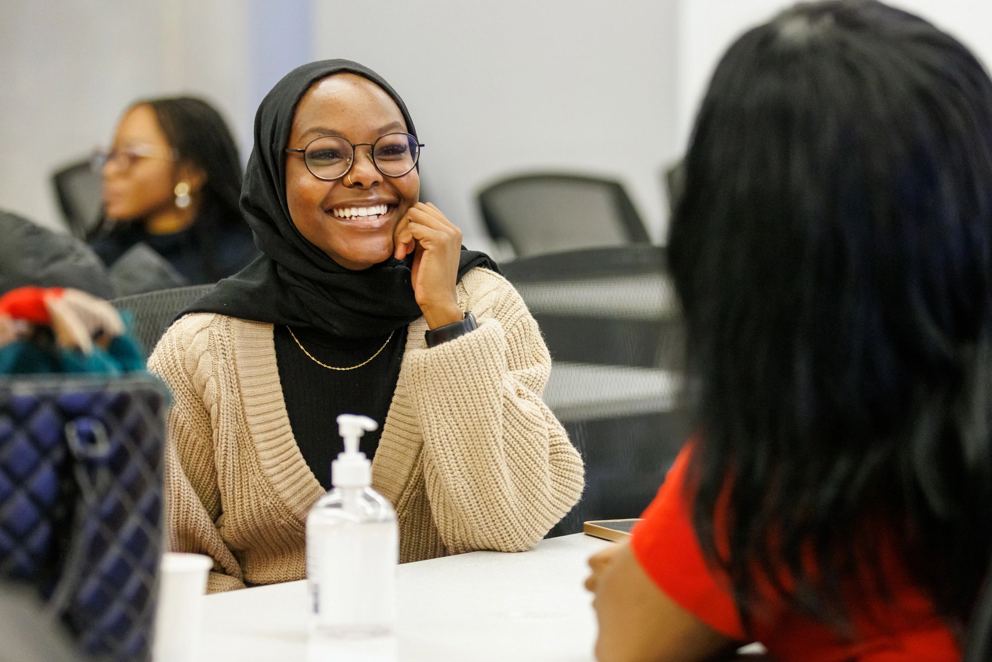 A student sitting at a desk smiles while in conversation with another person