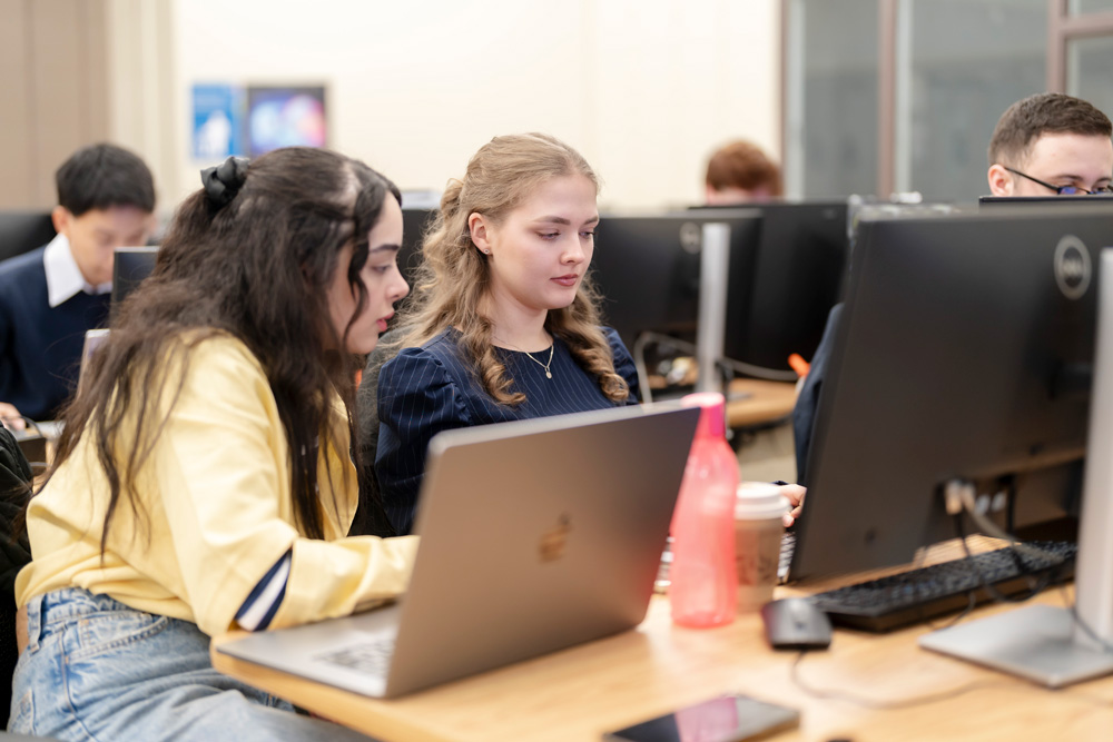 Two women seated in a classroom work at computers