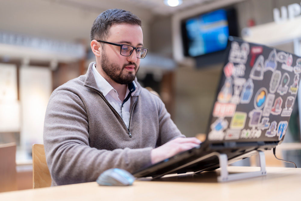 A beared man in glasses and a grey sweater types on a laptop