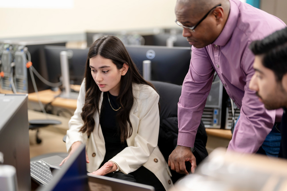 Two students work on their computers while a faculty provides direction