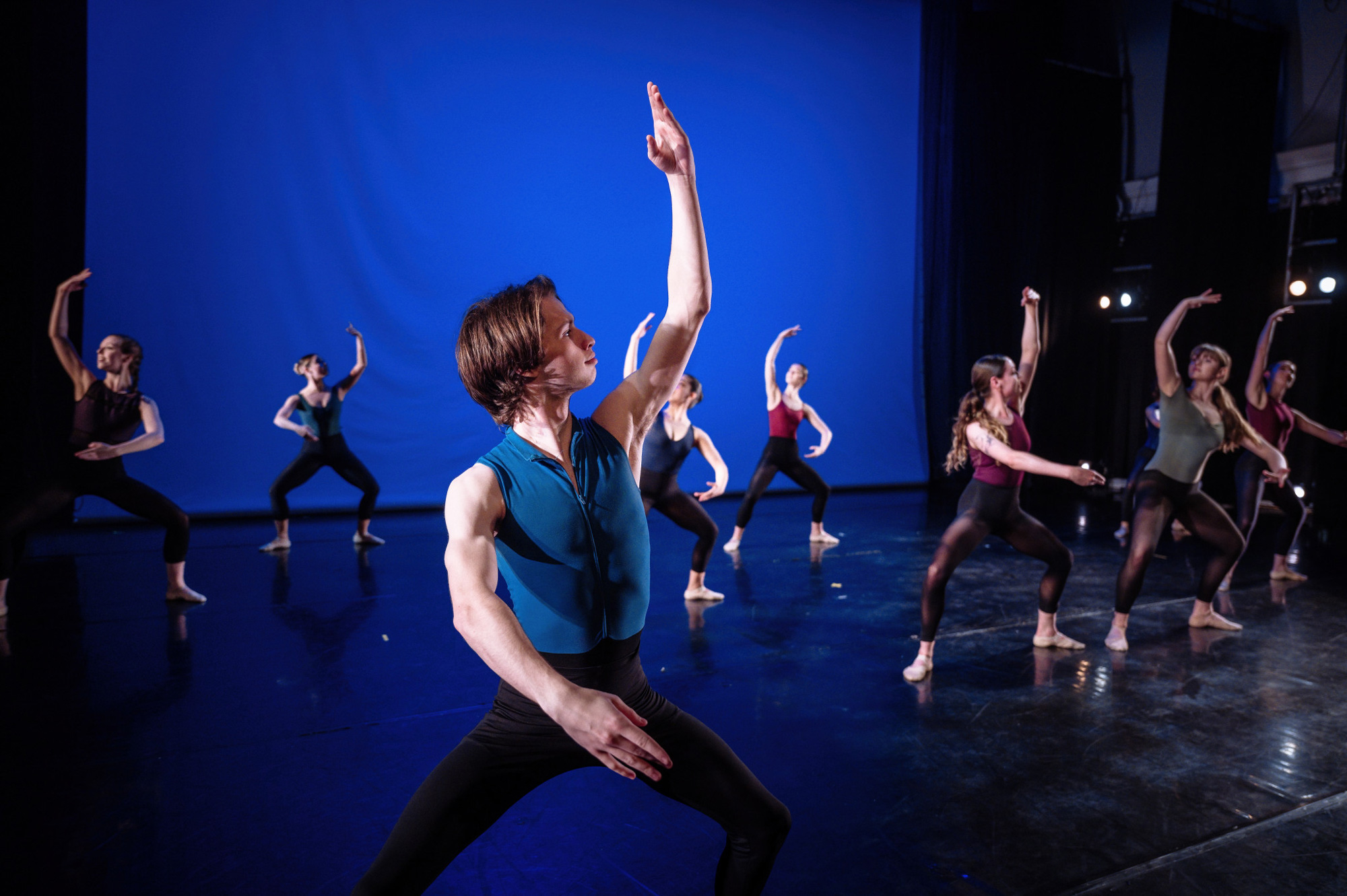 A dancer in a blue top and black tights strikes a pose on a dark stage with a blue background