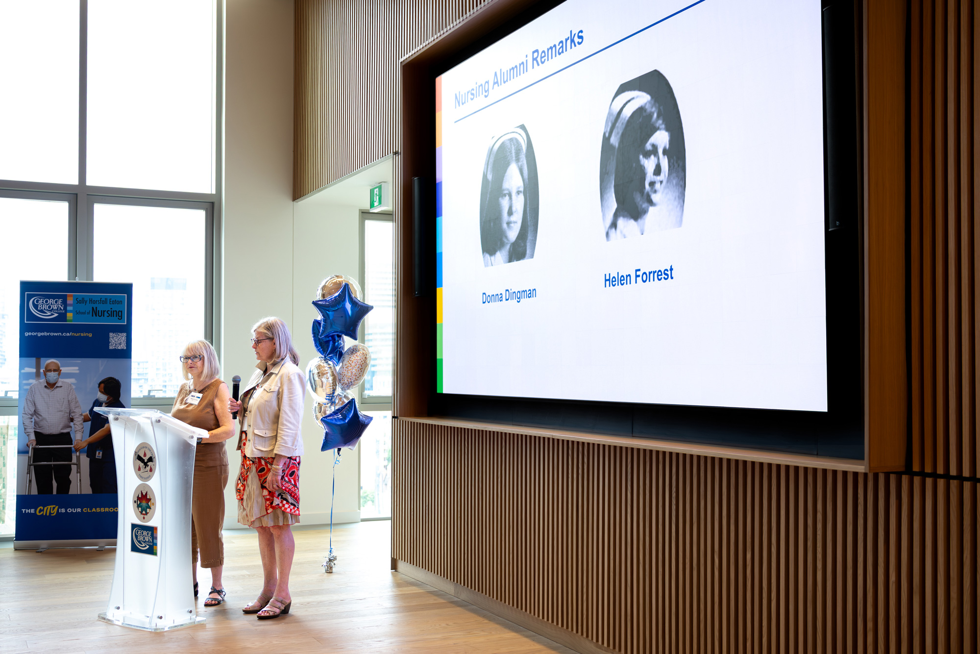 Donna Dingman and Helen Forrest speak to nursing alumni at a lectern in front of an image of themselves as nurse graduates in 1975