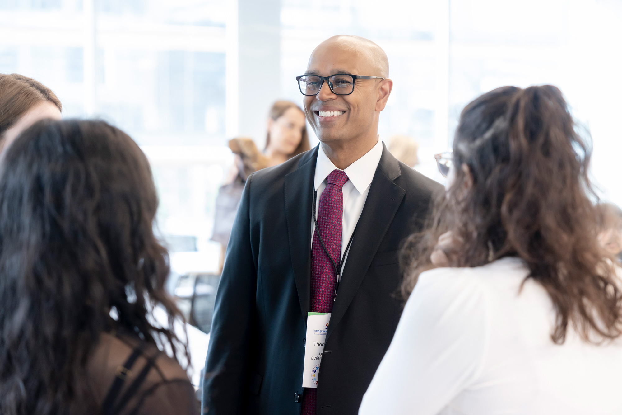 A man in a black jacket, red tie, and glasses smiles as he chats with a group of conference delegates