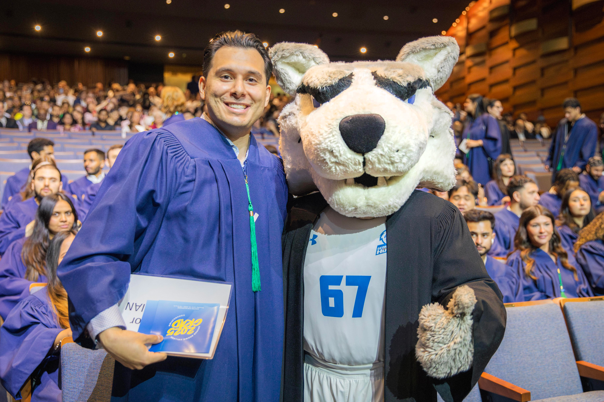 A student in a graduate robe poses with Helder the Husky in an auditorium full of people