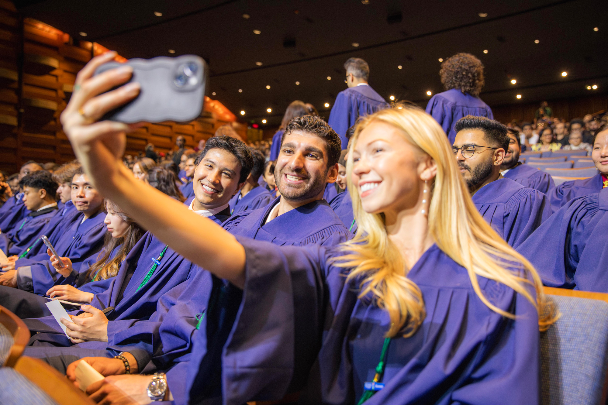 Seated students in their graduation gowns take a selfie together