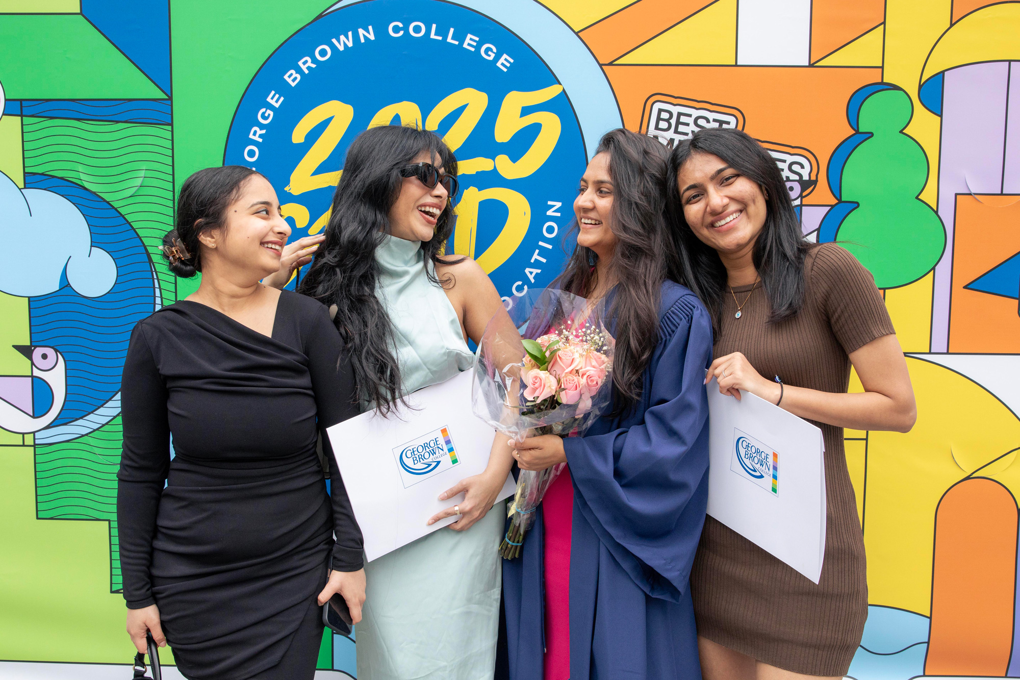 Four GBC graduates pose with their credentials following graduation in front a bright colourful mural