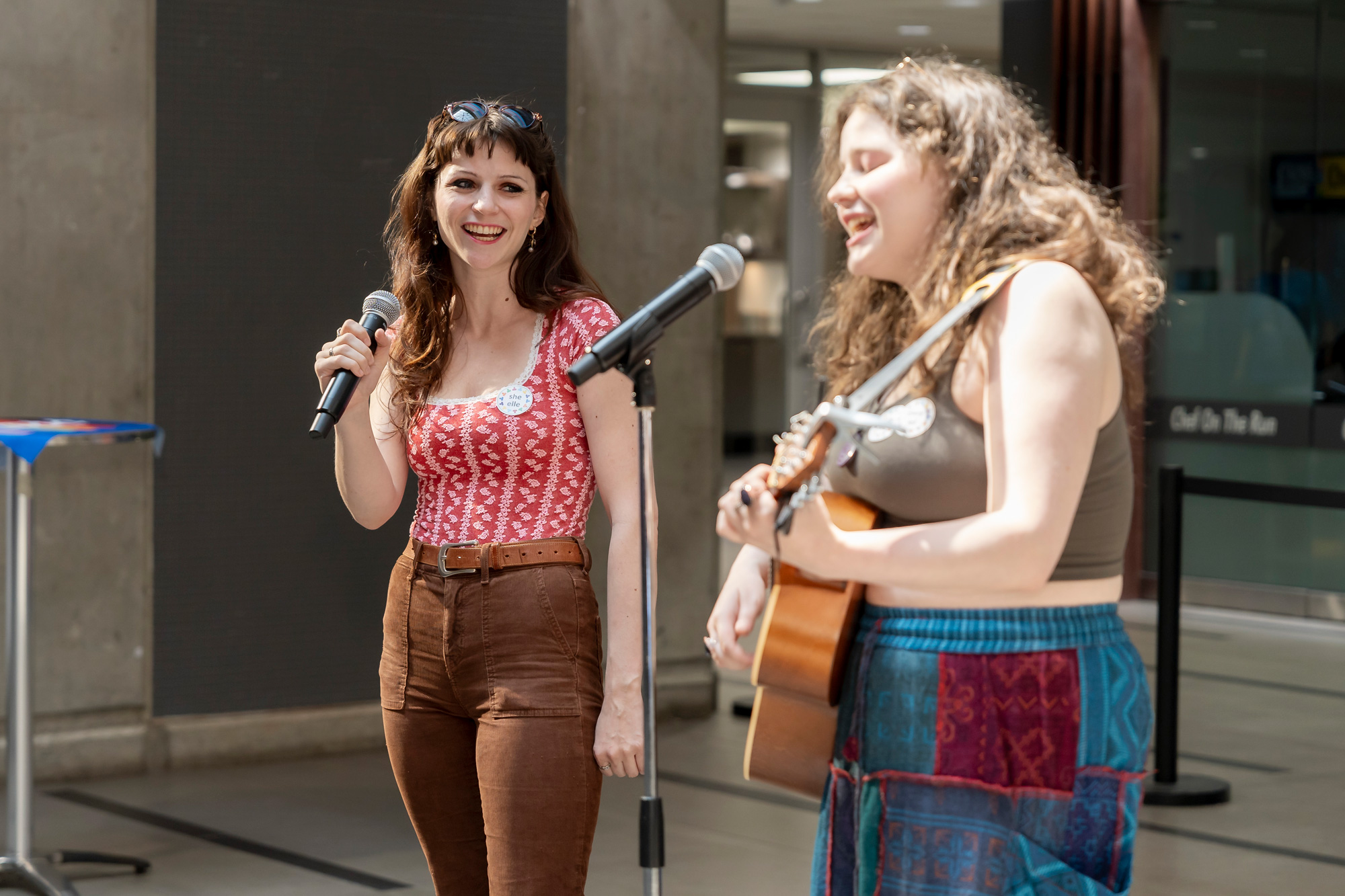 Two women perform, one holding a guitar, while both sing into microphones