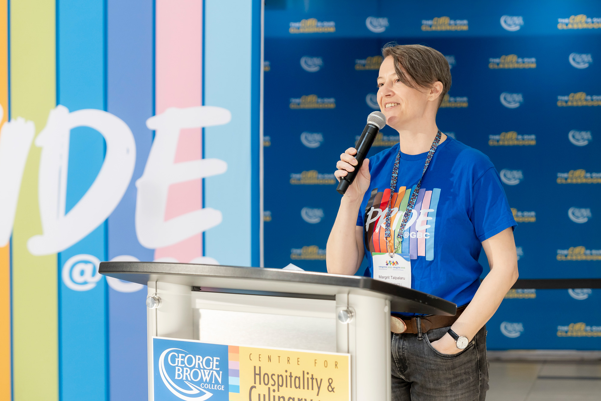 Dr. Margrit Talpalaru wears a blue shirt with Pride at GBC written on the front, holding a microphone at a podium