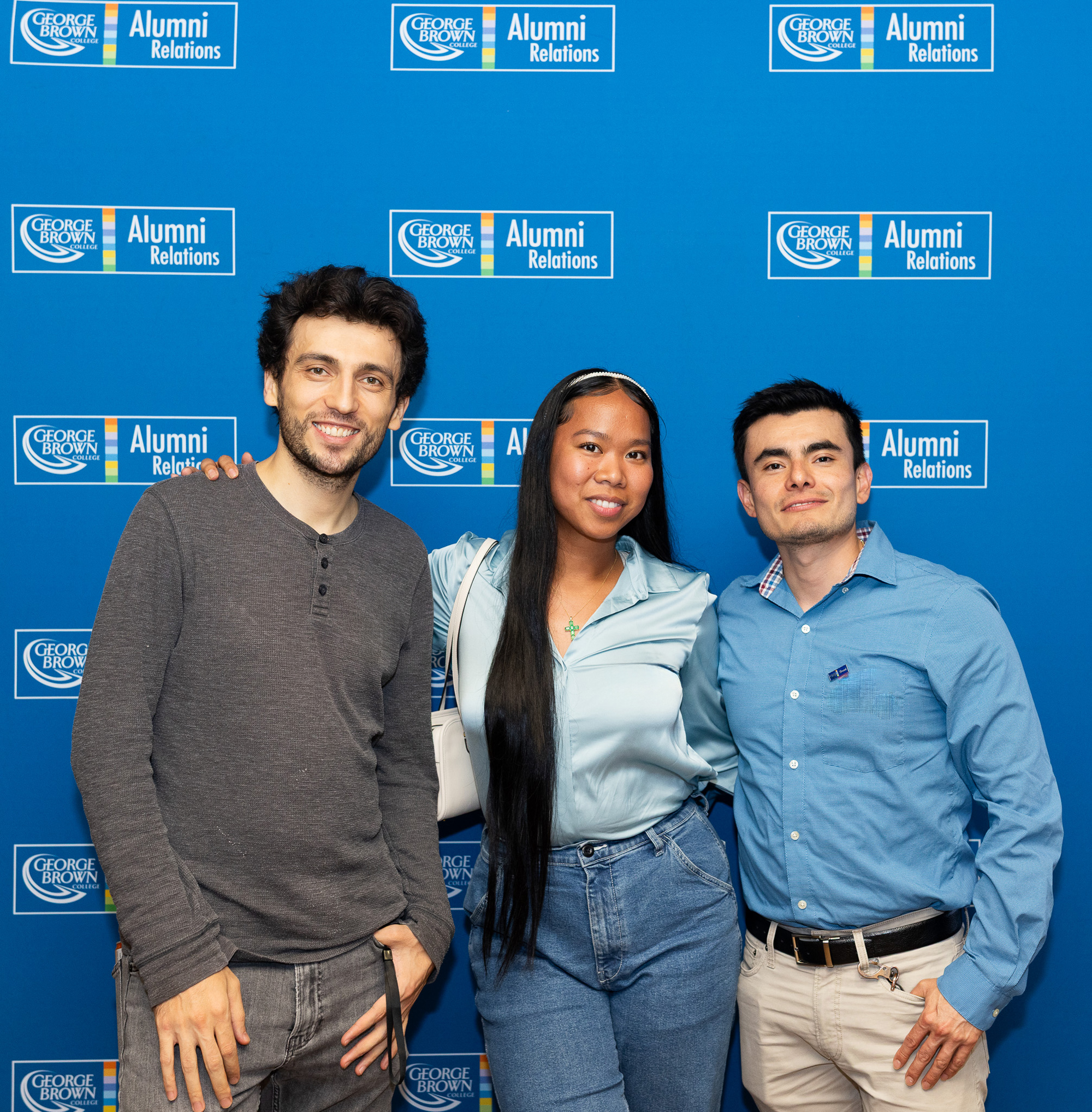 Three alumni pose in front of Alumni backdrop at the summer social event