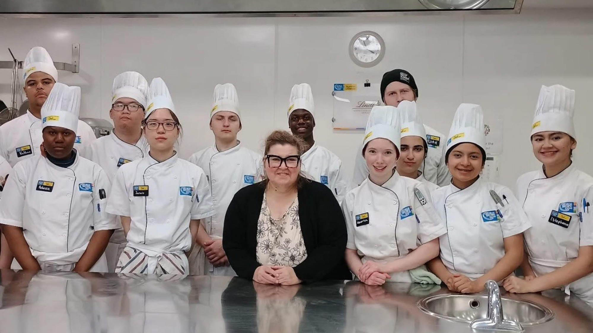 Brenda Torrens with Cook Level 1 students in chef uniforms during culinary training in kitchen classroom.