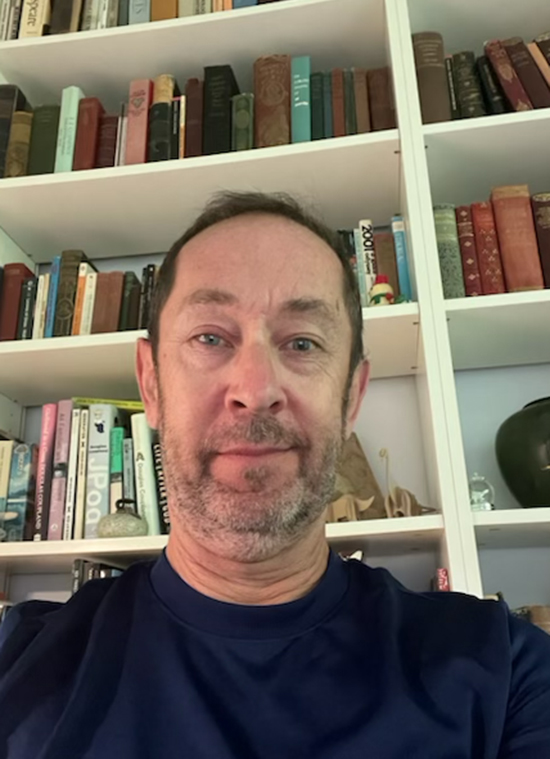 A man in a black t-shirt with facial hair in front of a bookshelf