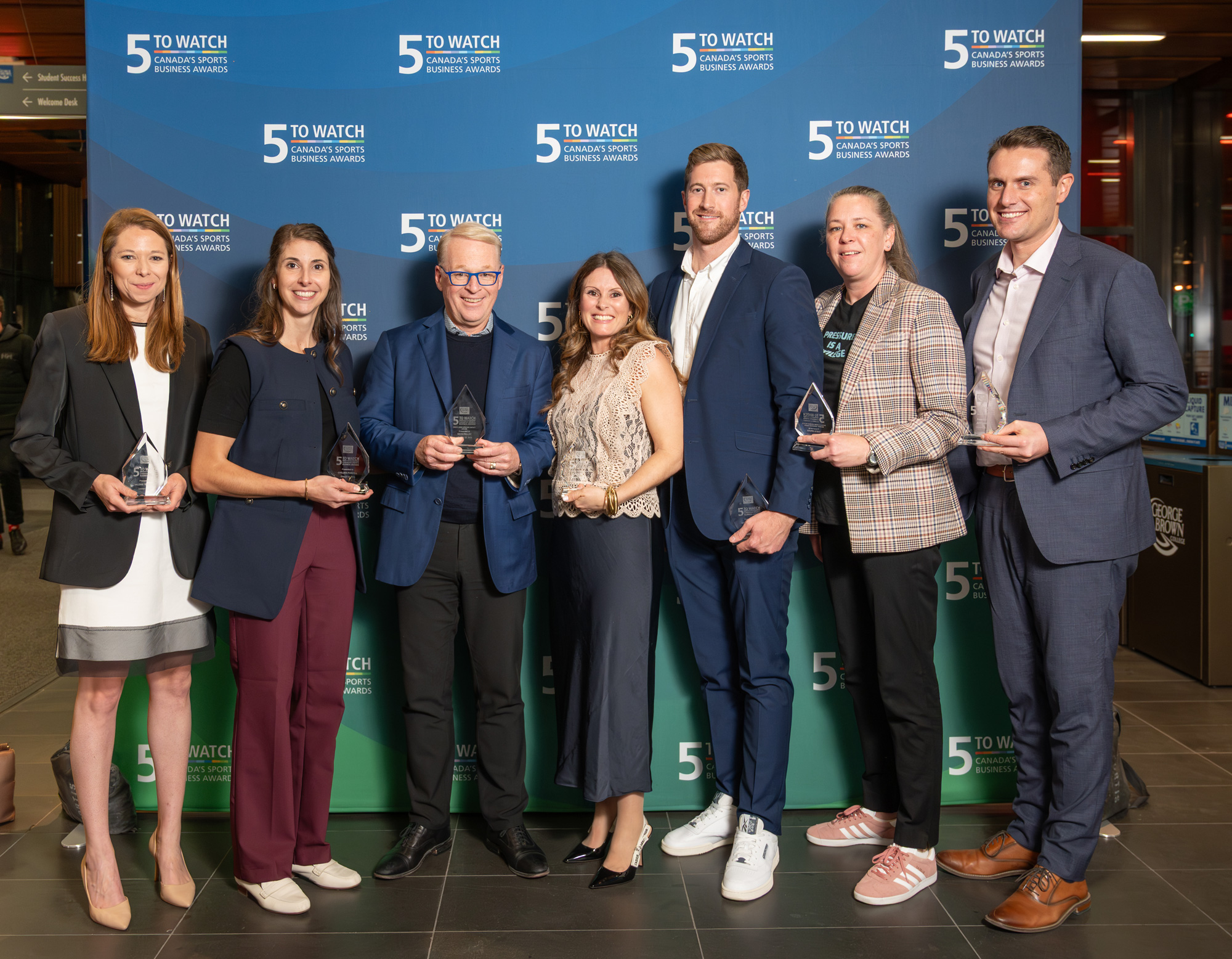 2025 5 to Watch award recipients pose together with their awards in front of a blue background
