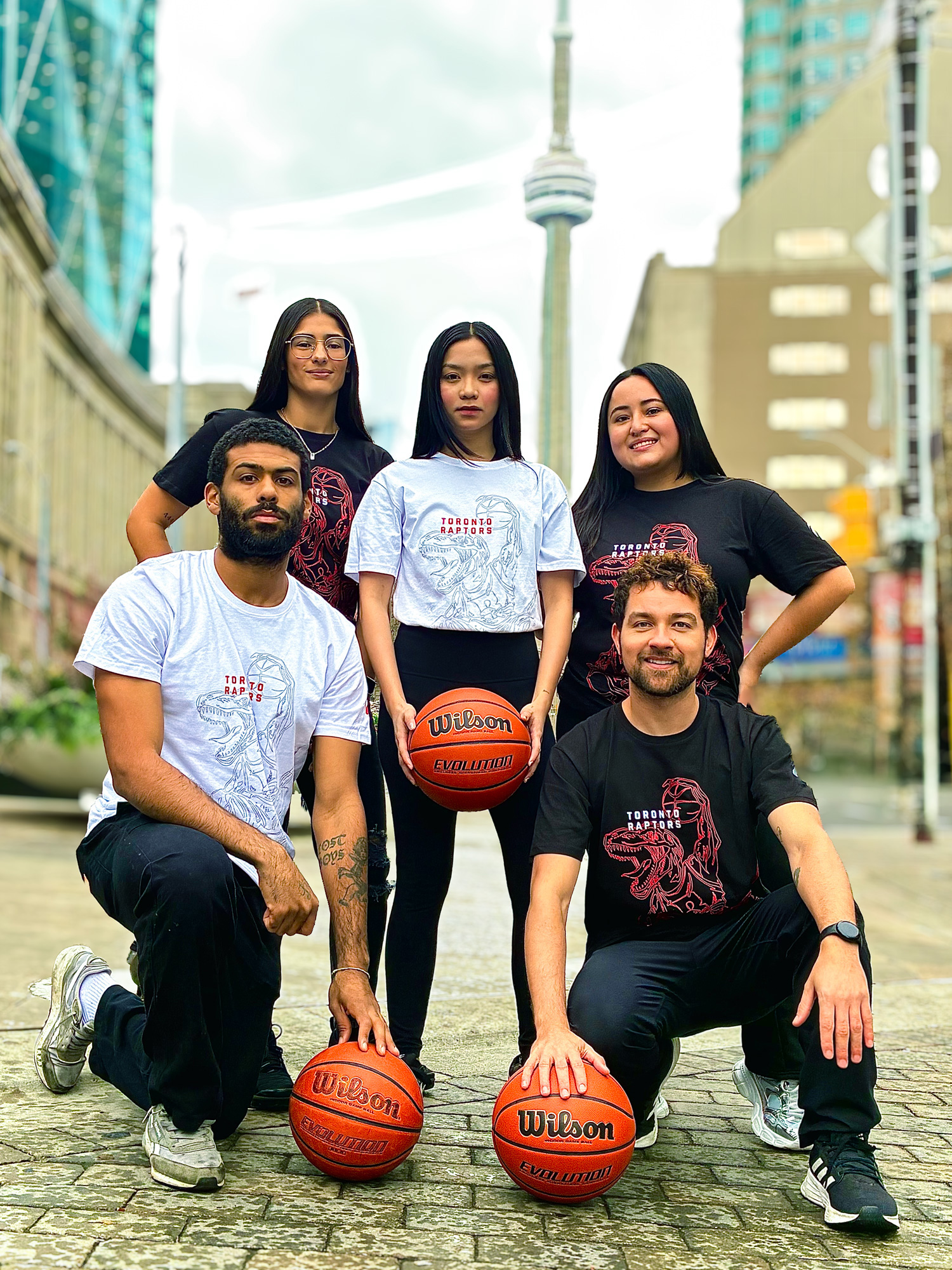 A group of five students pose on the street in black and white Raptors t-shirts and basketballs with the CN tower behind them