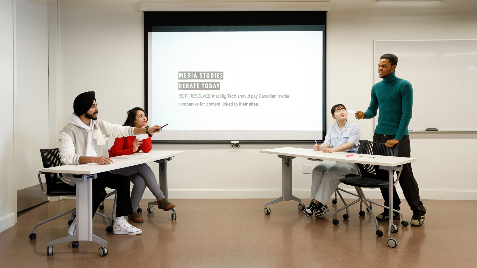 Students hold a debate on Media Studies. There are two sides to the debate with the students sitting at tables opposite each other while one student stands and has the floor.