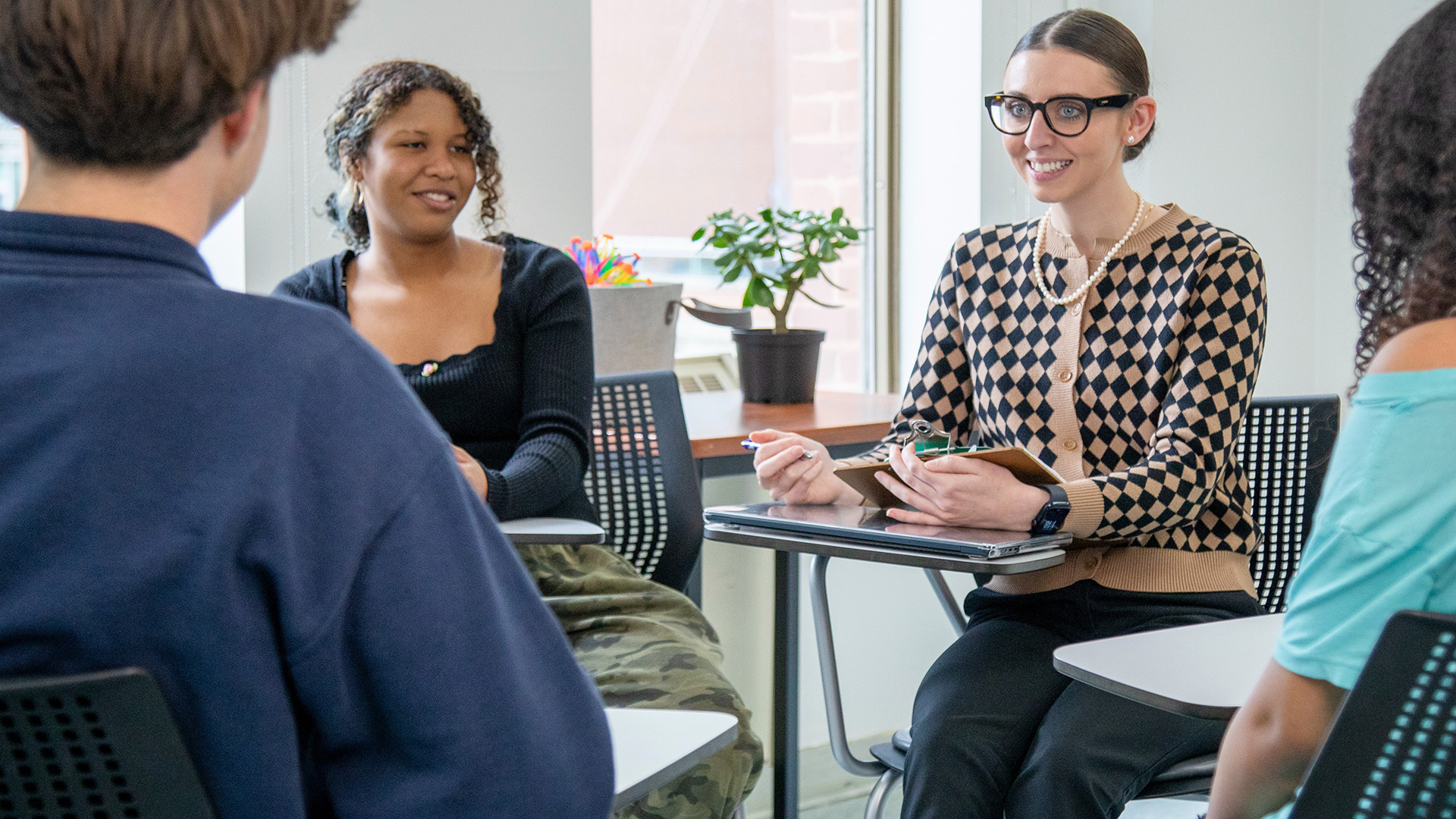 Instructor and students in a child and youth care program sitting in a small classroom circle, engaged in a supportive group discussion, with the instructor holding a clipboard and smiling as students share.