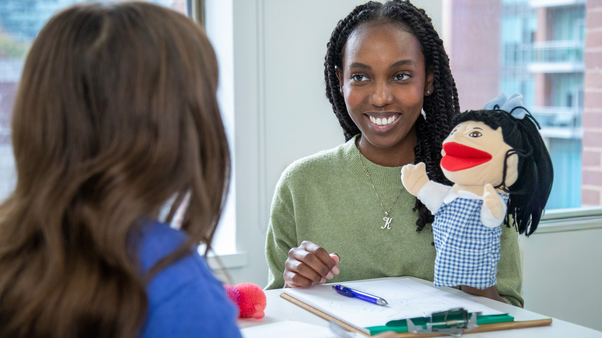 Student in a child and youth care program using a hand puppet during a practice session, smiling as she engages with a peer in a mock therapeutic activity at a classroom table.
