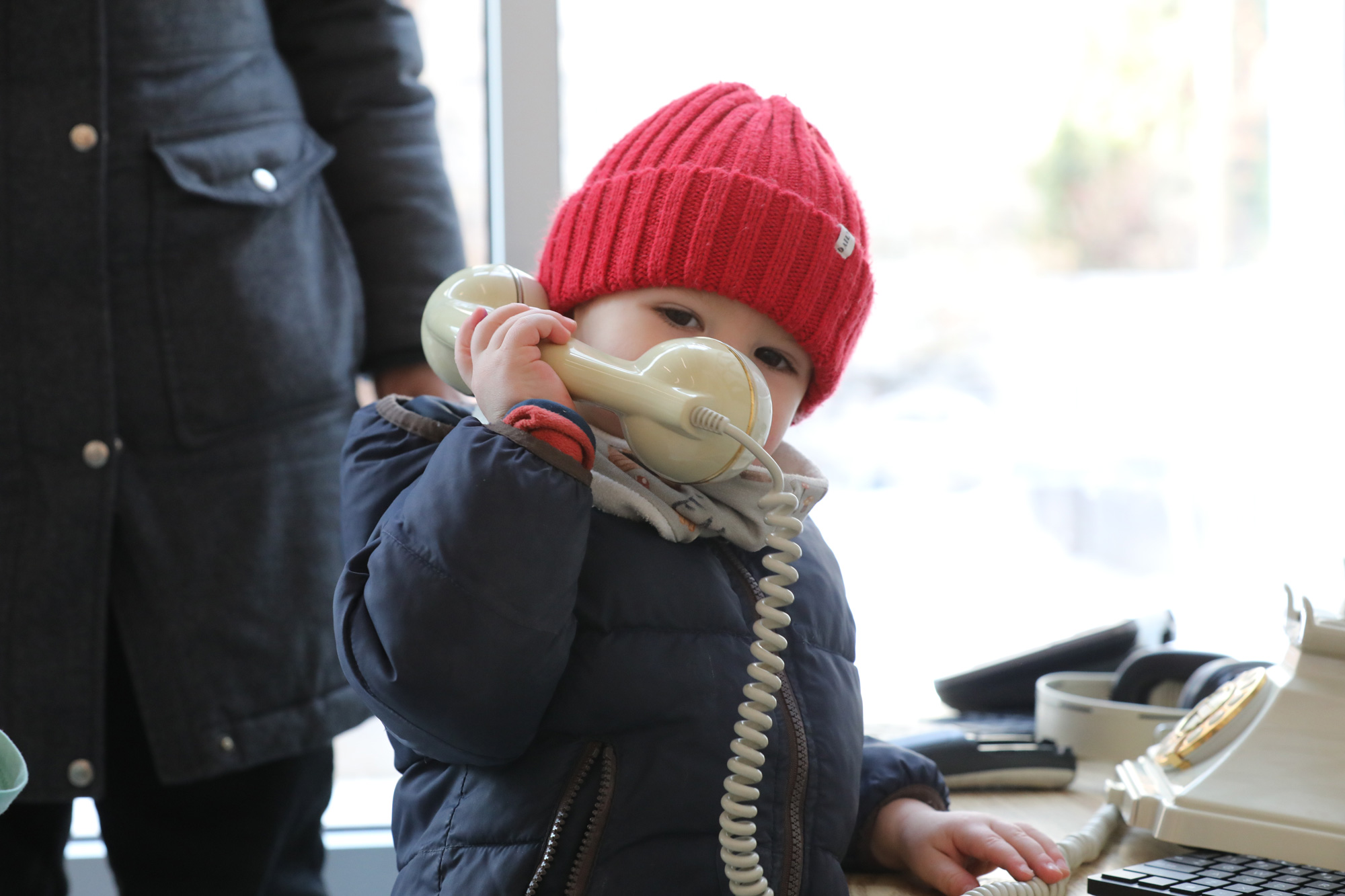 A toddler in a red hat holds up a telephone