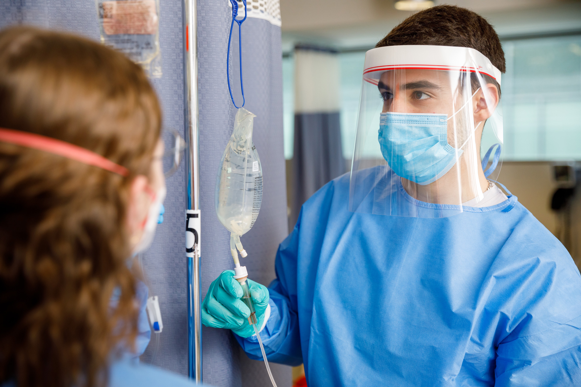 Healthcare professional in protective gear adjusting an IV bag while interacting with another individual in a clinical setting.