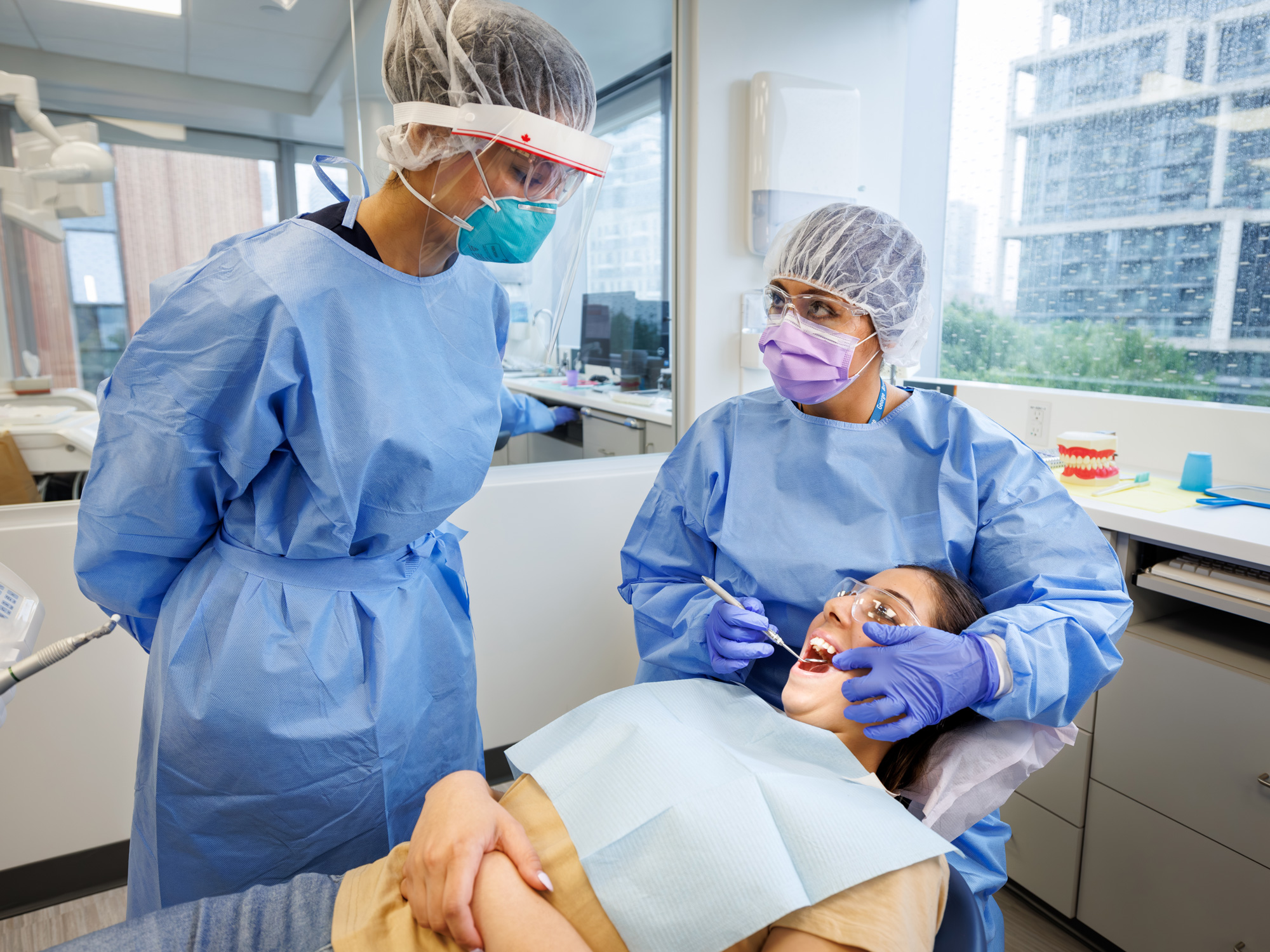 George Brown Polytechnic dental assistant students in a clinic setting performing an oral exam on a patient while wearing protective equipment.