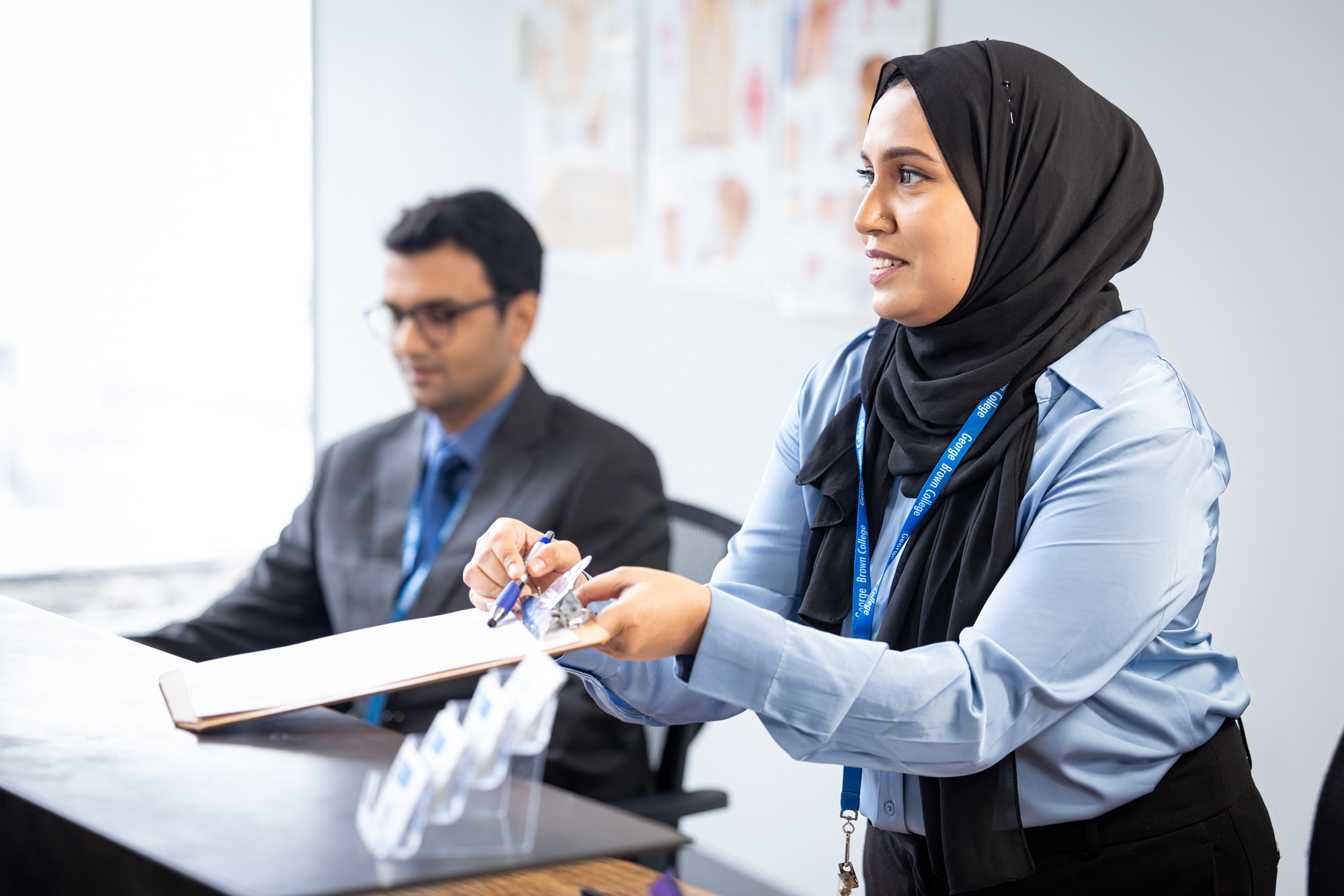Person at a George Brown Polytechnic reception desk holding a clipboard and pen while assisting a visitor, with another staff member seated nearby.