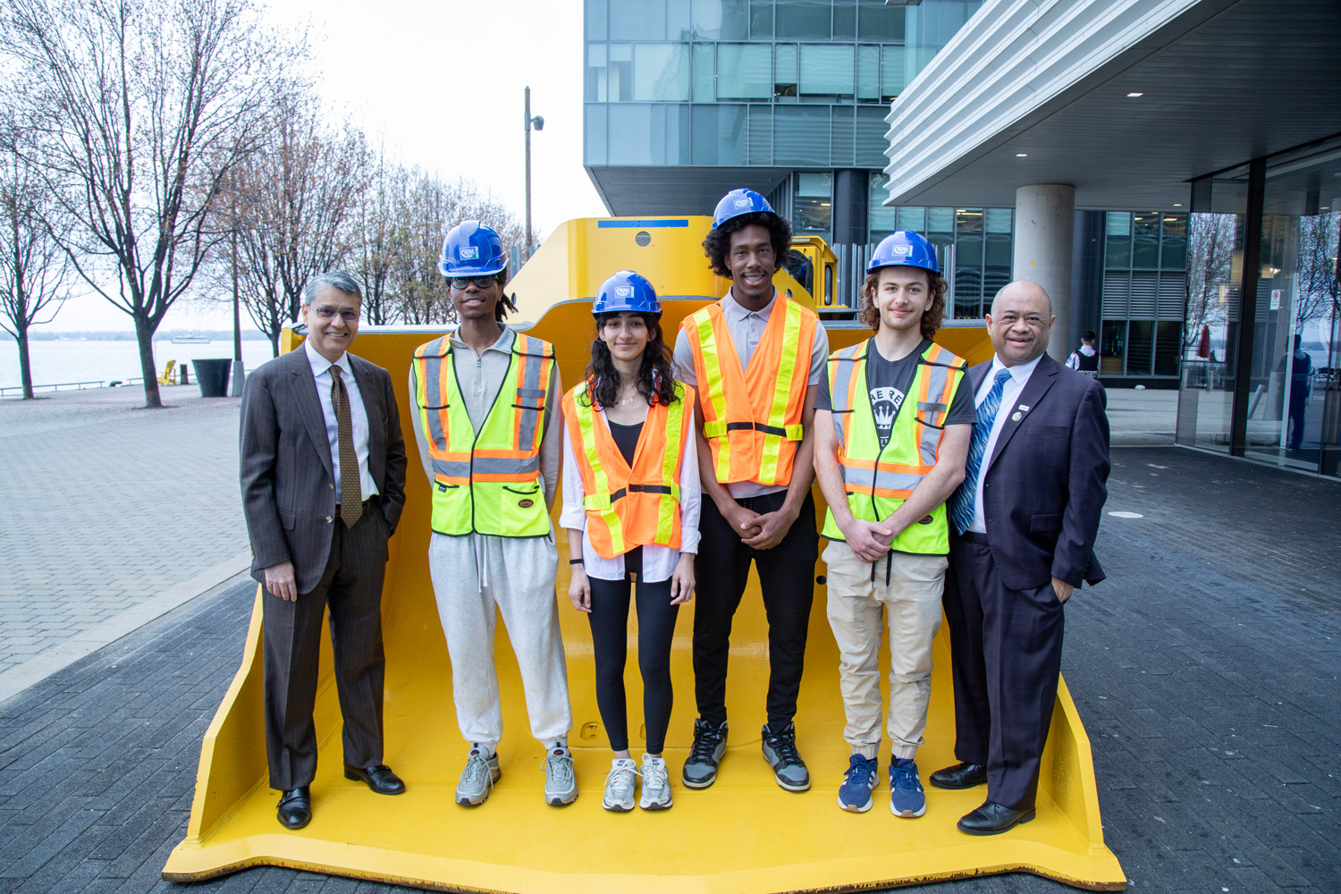 Dr. Hassun Malik and Micheal Herrera pose with students in hard hats and vests in the bucket of a digger at the Waterfront Campus
