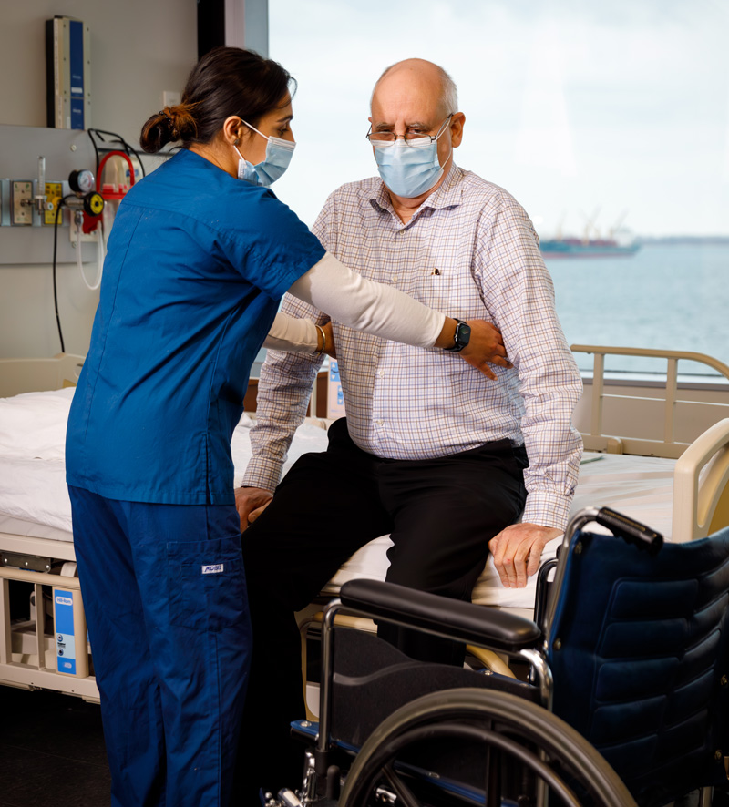 George Brown Polytechnic personal support worker student assisting a patient seated on a hospital bed during a care assessment, with a wheelchair nearby.