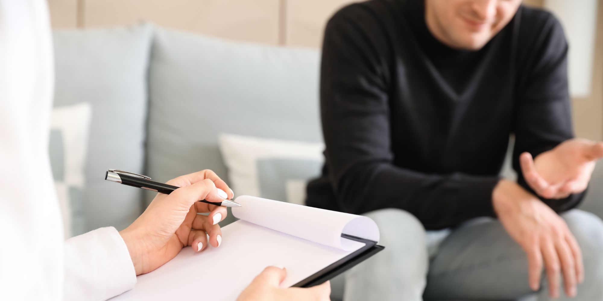 A close up of a counselling appointment. In the foreground, someone is taking notes. In the background, someone is sitting on a couch, gesturing with their hands while talking.
