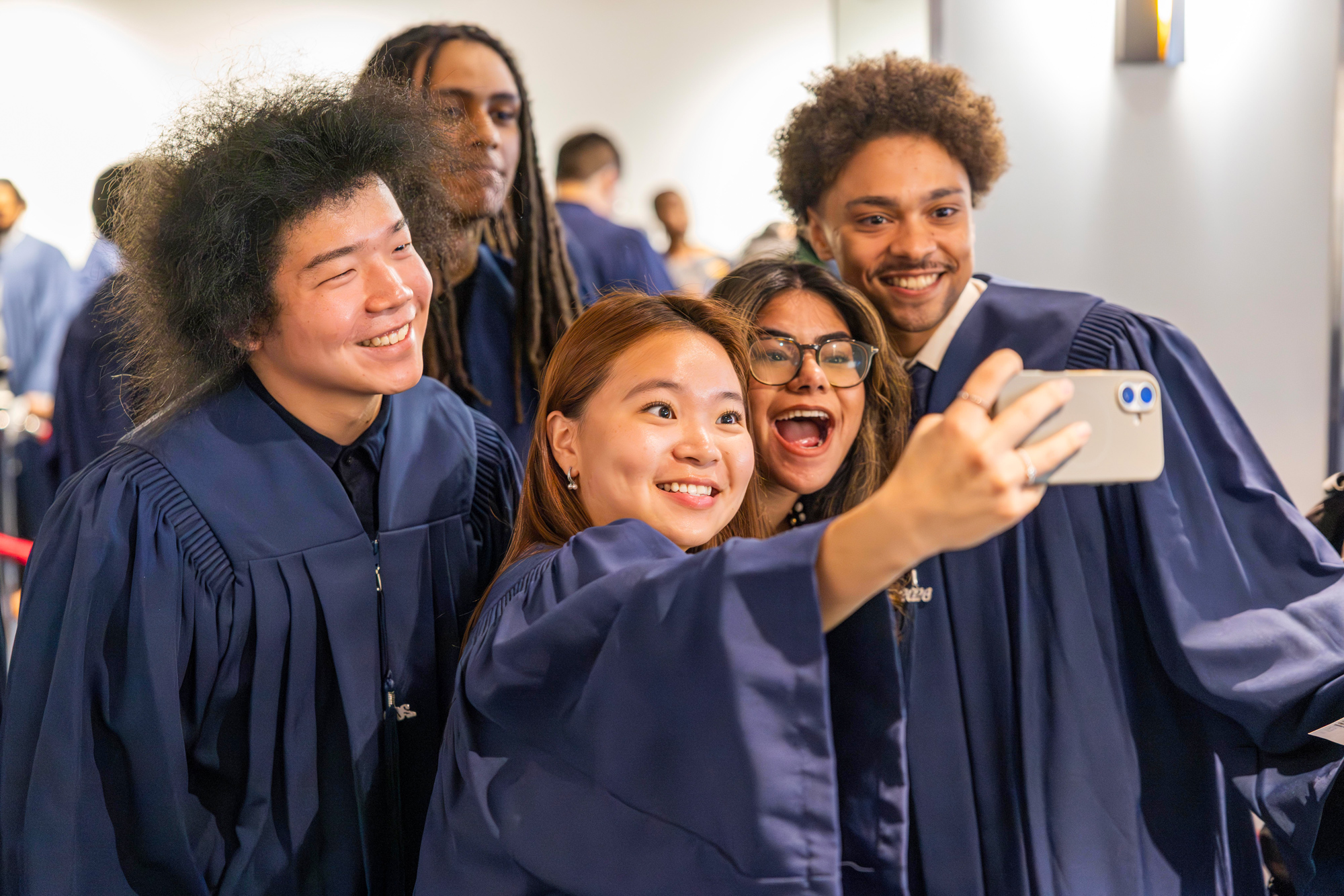 Five students in navy convocation gowns pose together and take a selfie.