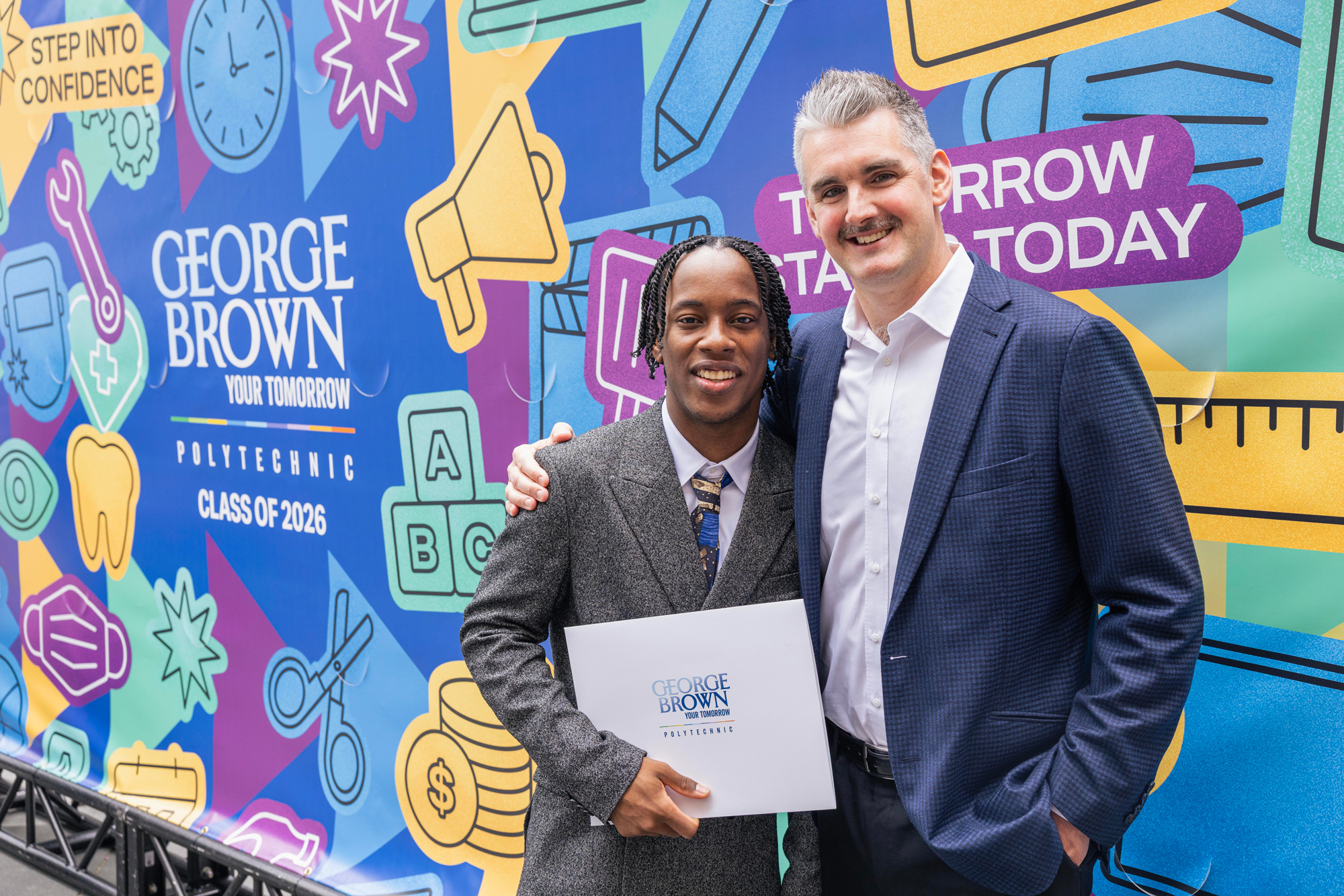Negus King holds up his credential with his chef mentor in front of the Convocation mural.