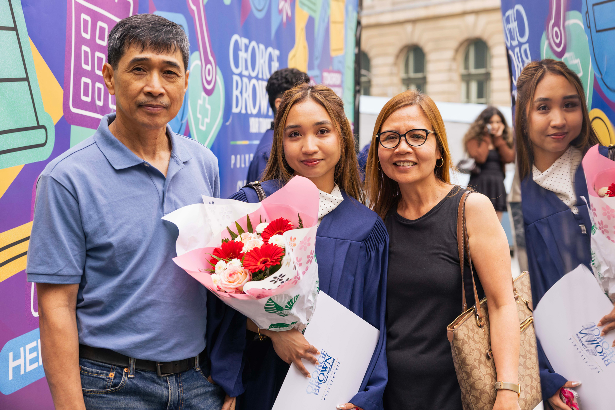 A graduating student holds flowers in their gown with family surrounding them against a bright colourful background.