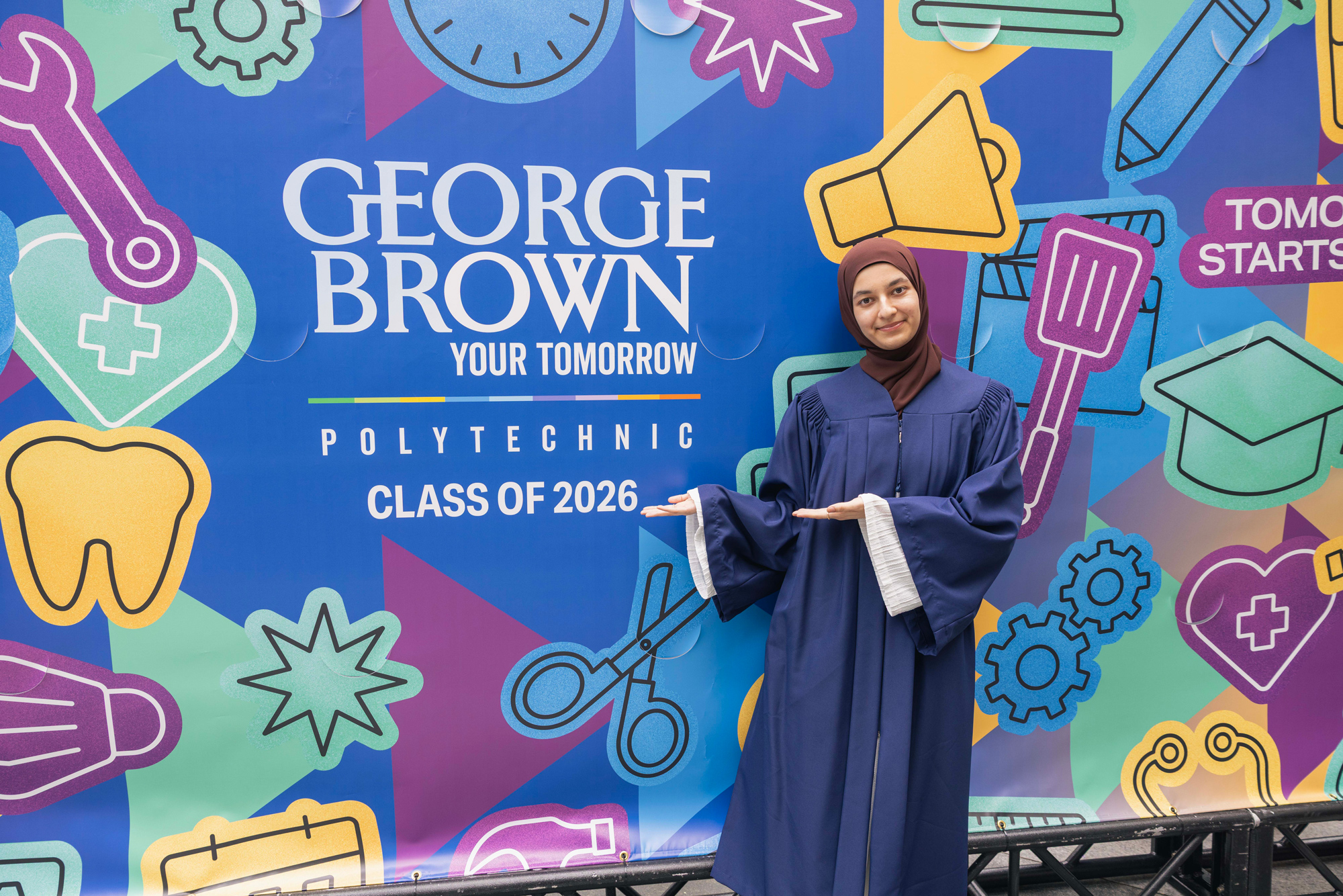 Madina Amanzai stands in a her convocation gown in front of the bright and colourful convocation mural she design for the Class of 2026.