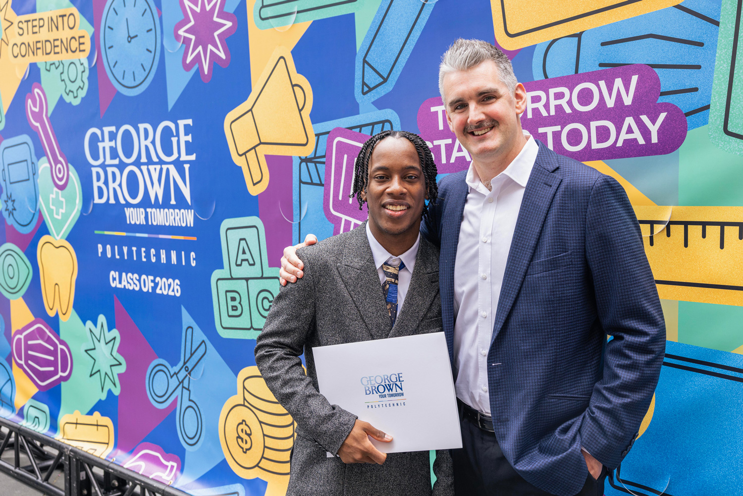 Negus King holds up his credential with his chef mentor in front of the Convocation mural.