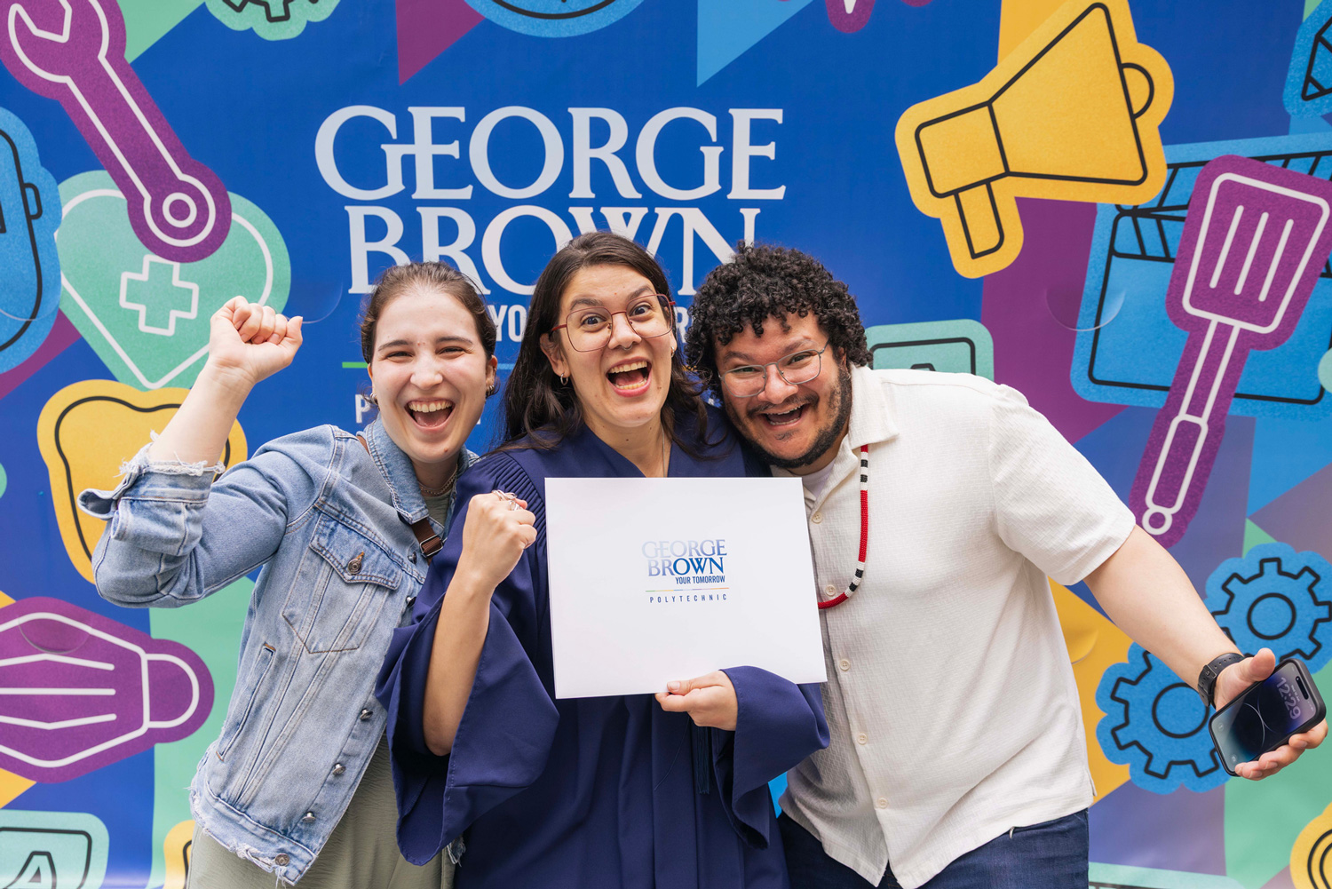 A new grad cheers with friends holding their new credential against a bright and colourful mural.