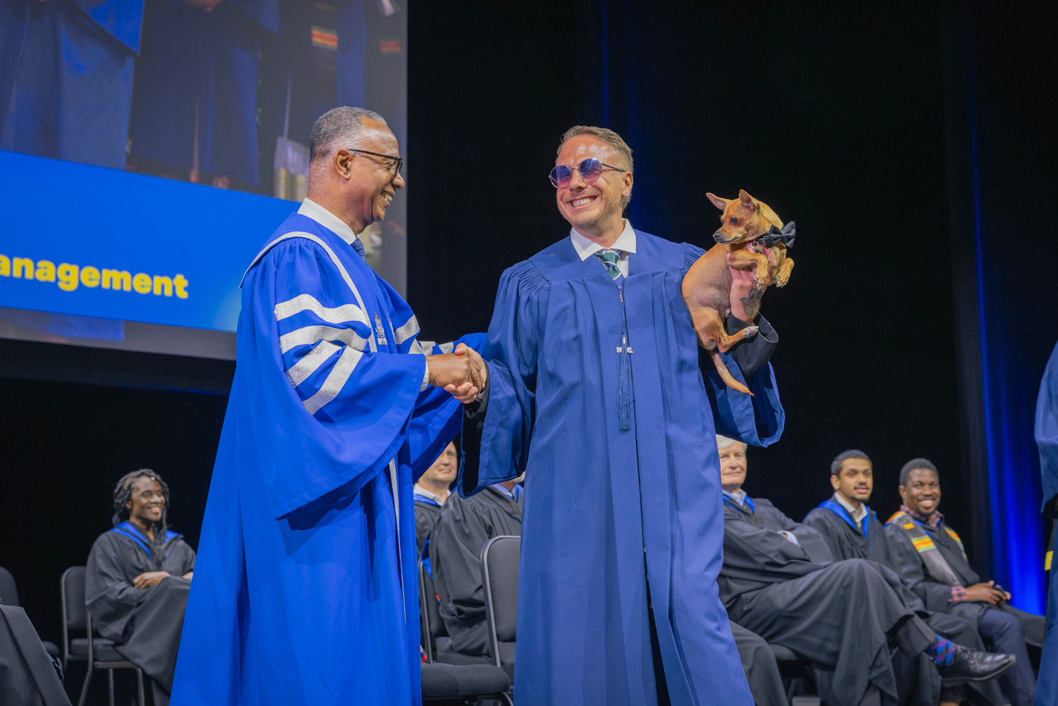 A graduate with glasses holds a small dog as he crosses the stage and shakes Dr. Fearon's hand at Convocation.