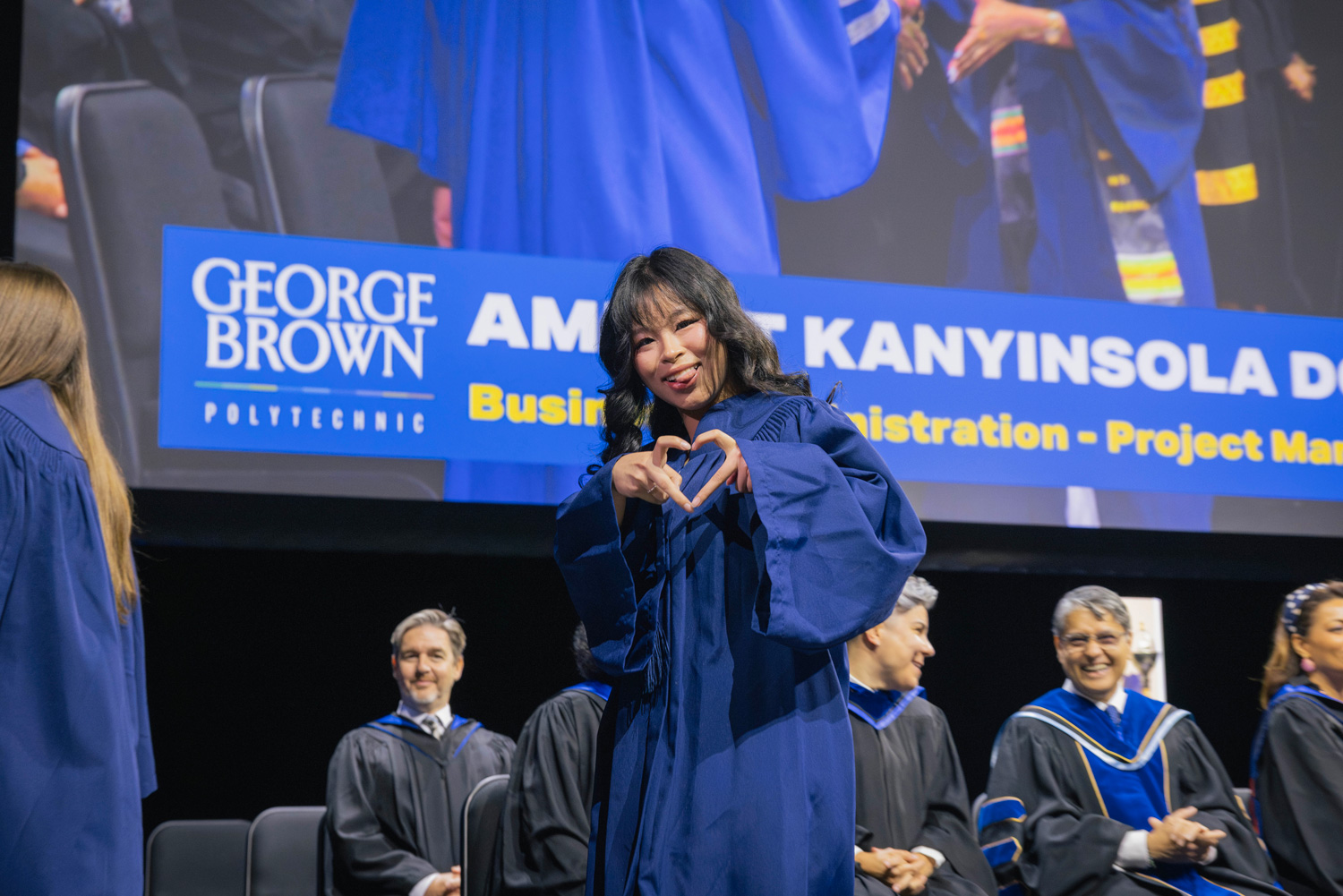 A grad makes a heart shape with her fingers as she crosses the stage at convocation.
