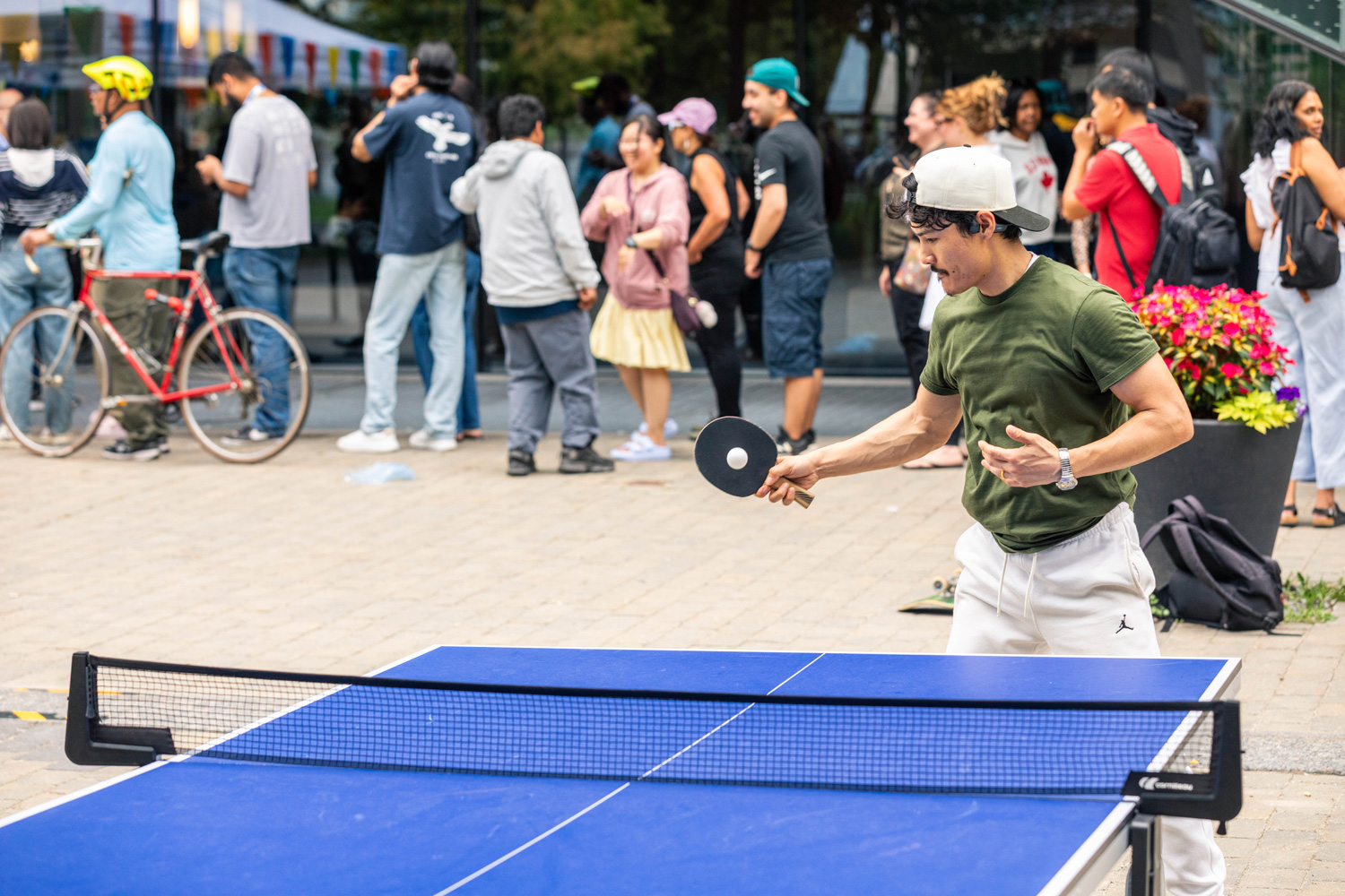 Students play table tennis outdoors while guests and tents with bunting are seen in the background.