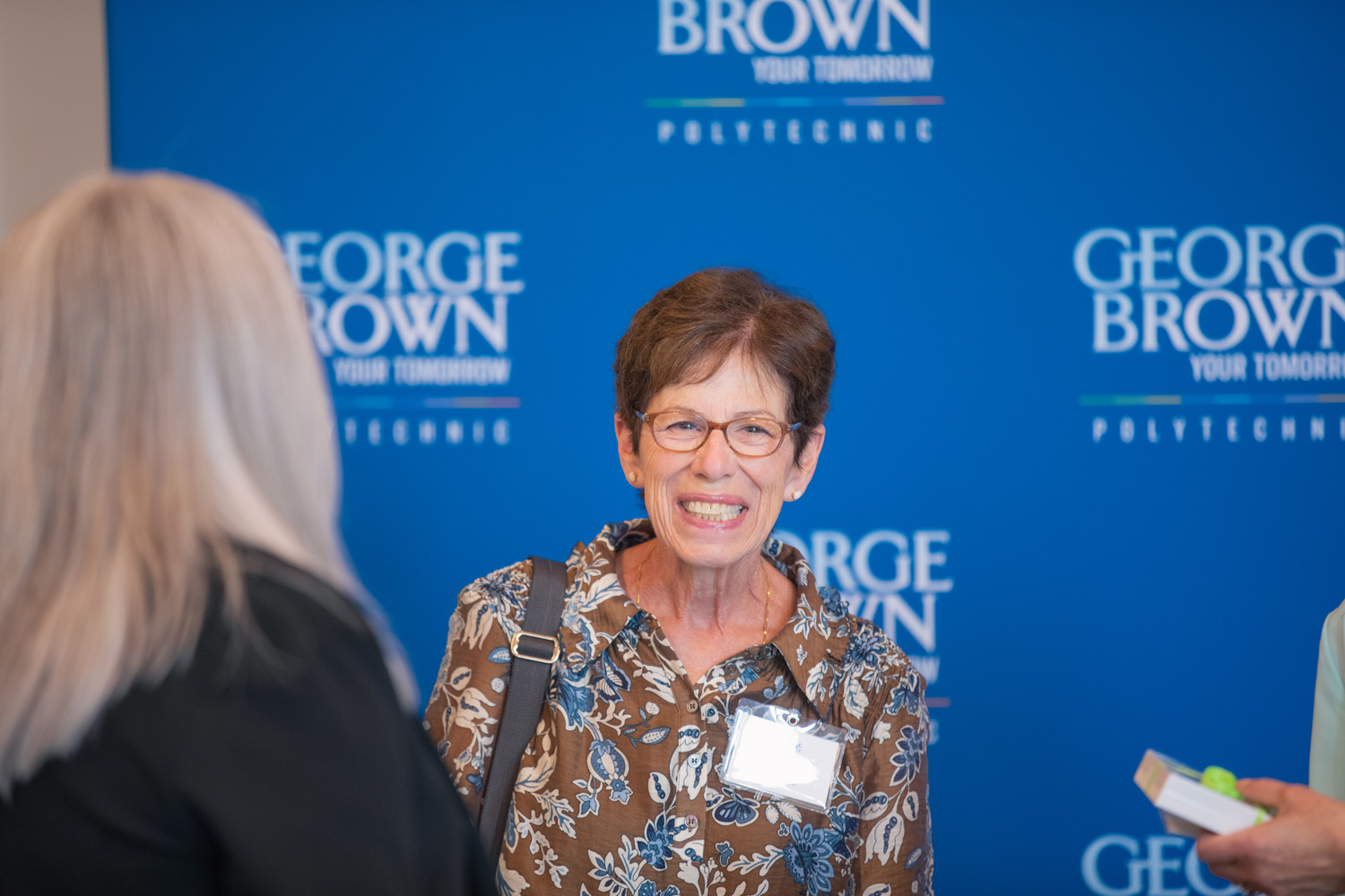 A 1976 nursing grad smiles for the camera in a floral shirt in front of a blue George Brown background.