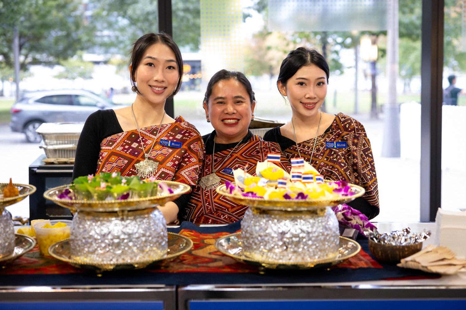 Three women in South East Asian clothing stand behind colourful plates of food.