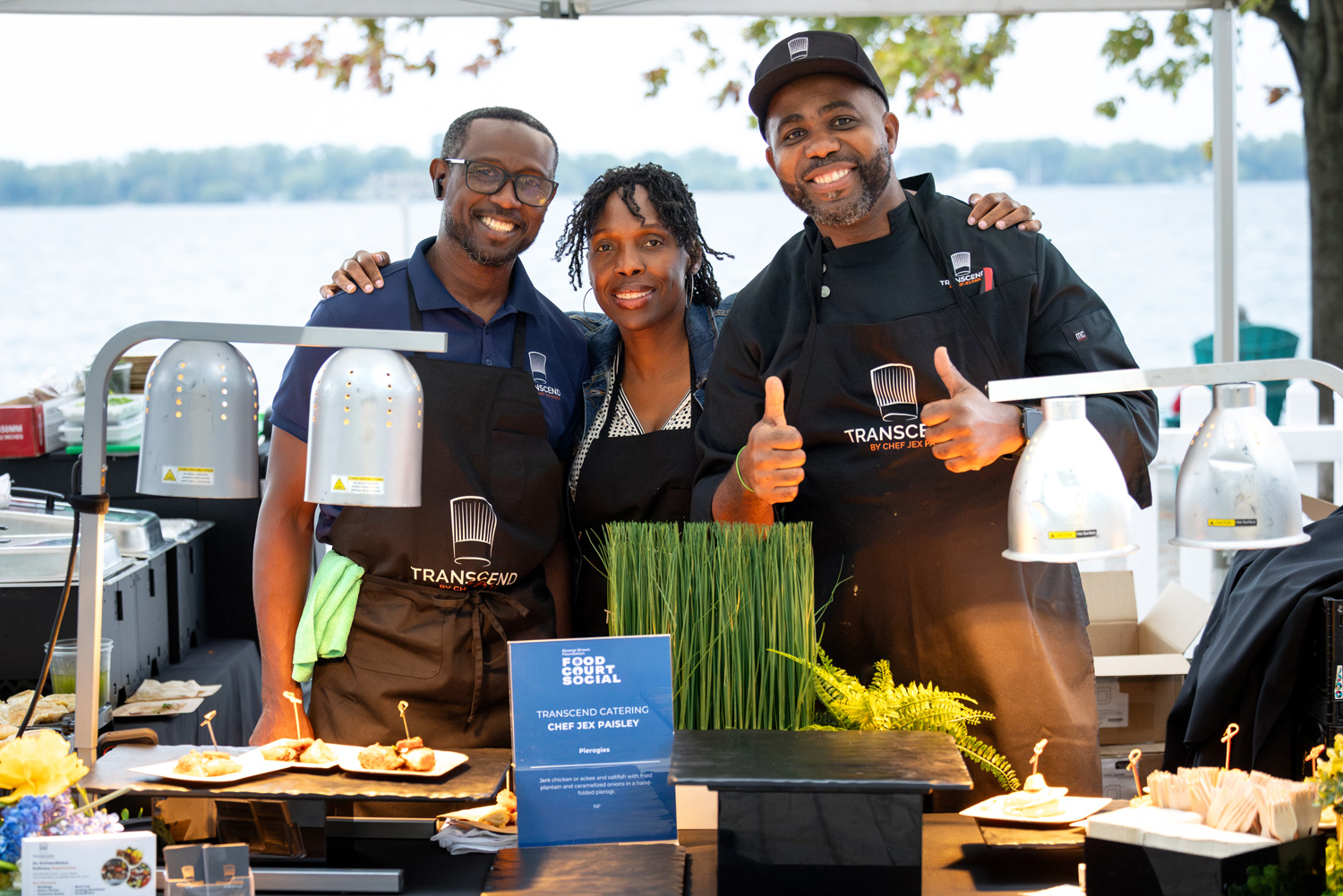 Three chefs in black aprons from Transcend Catering smile behind their table with a view of the lake behind them at the Food Court Social.