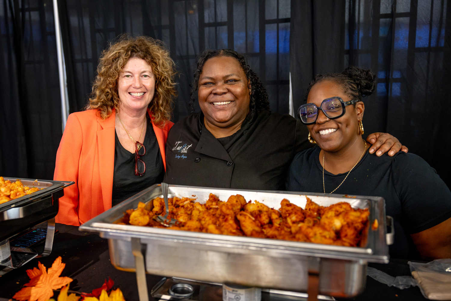 Three women stand behind trays of hot wings and southern food at the Food Court Social.