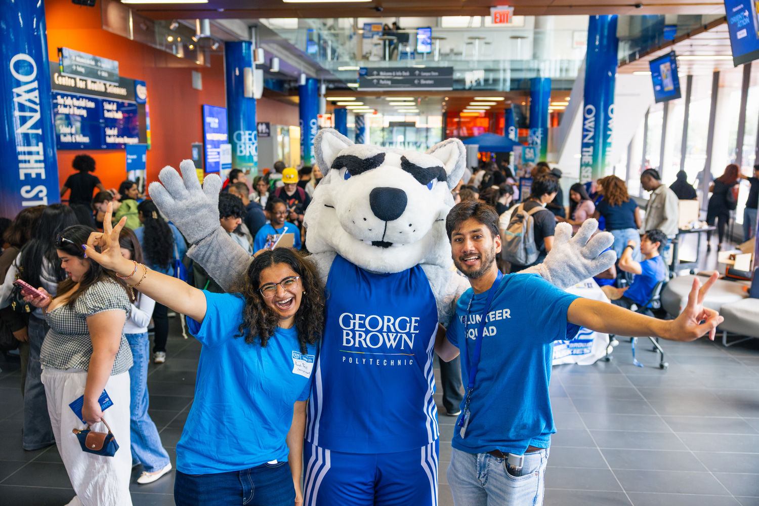 Two students pose with arms outstretched with Helder the Husky at Waterfront Campus.