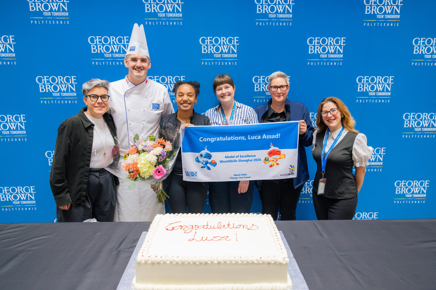 Luca and the Chef School team hold up a banner welcoming her back with a cake that reads 'congratulations Luca'.