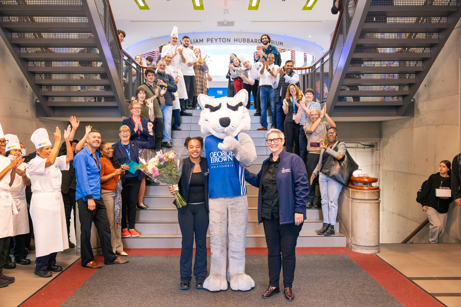 Staff, faculty, and students line the stairs to the Chef School welcoming Luca back from her competition in Shanghai. Luca, Helder the Husky, and Associate Dean Tara Ferguson stand in the middle smiling.