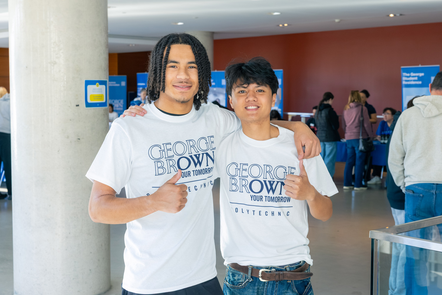 Two students in white George Brown Your Tomorrow t shirts smile with thumbs up.