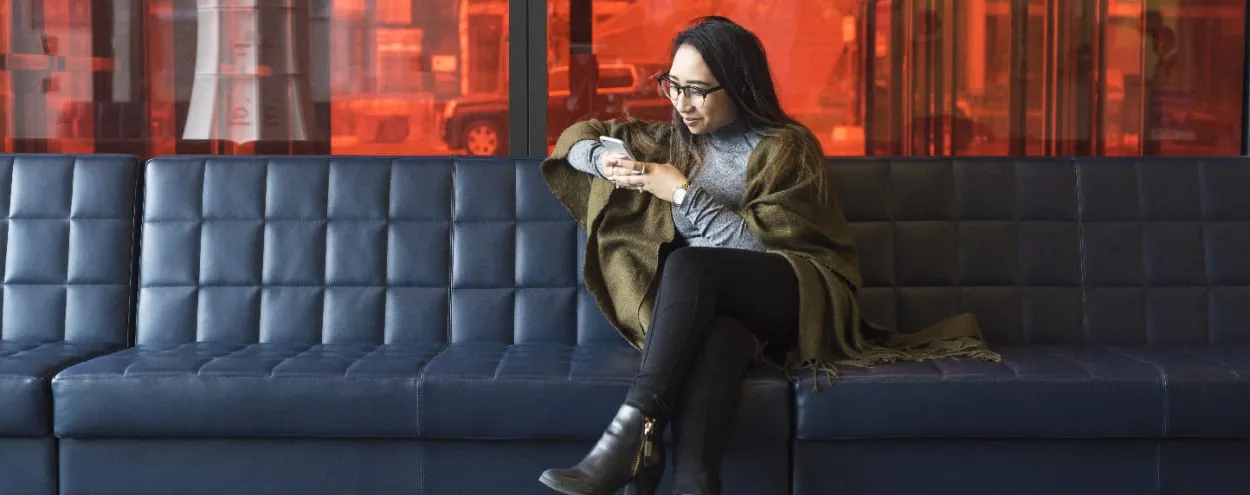 A female student is sitting casually on a comfy lounge bench at the Waterfront Campus and looking at her phone or texting to someone while smiling. 