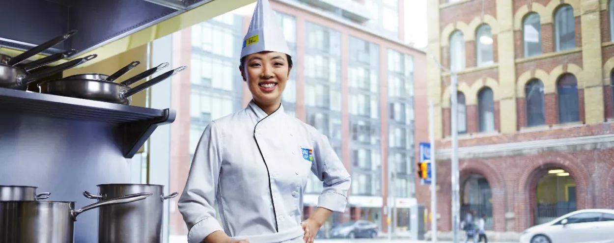 Female Chef School student in uniform poses by stove and window in kitchen/classroom.