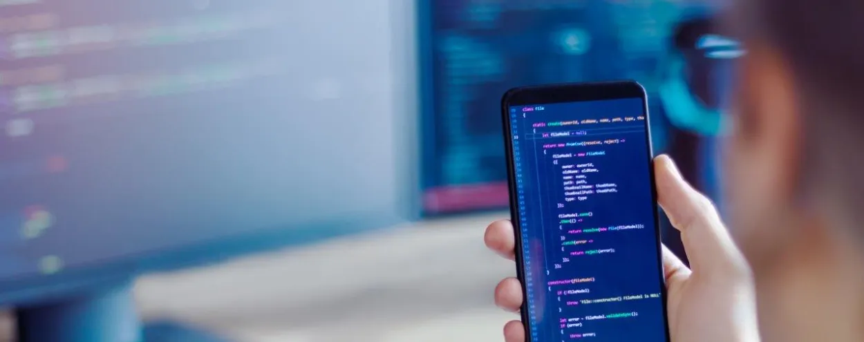 A hand holding a mobile phone with computer code on it in the foreground with a desktop computer in the background.